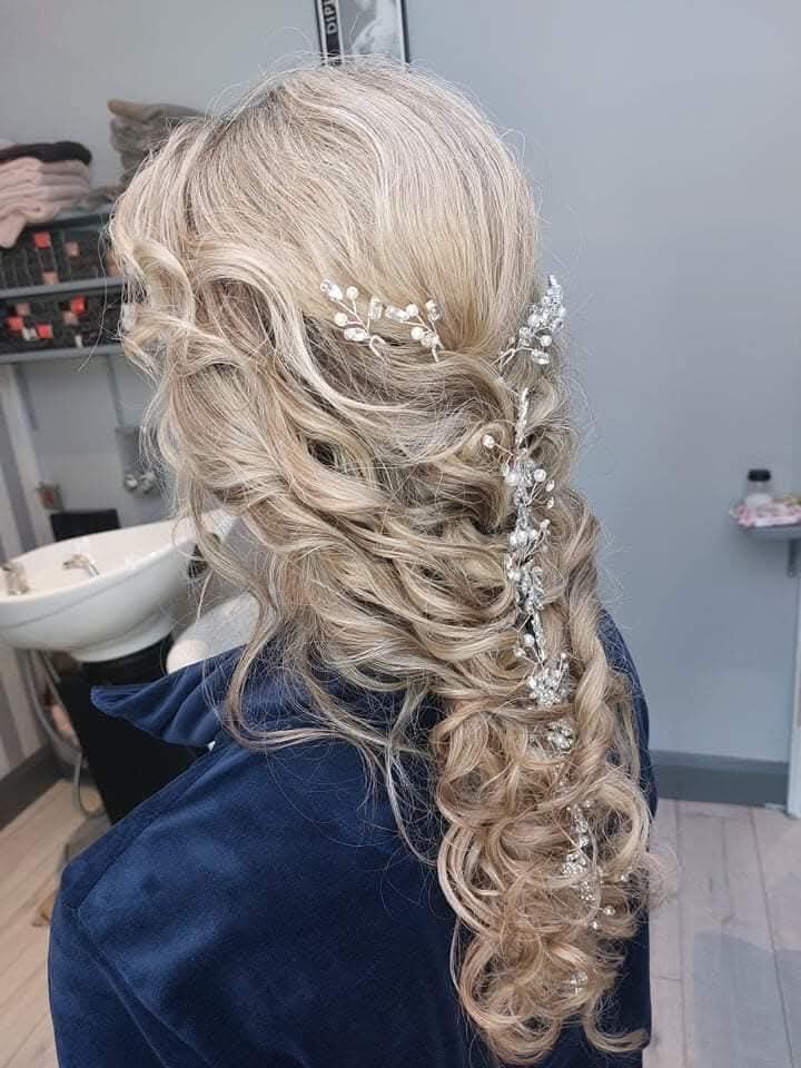 Bridal half-up style on long blonde hair: loose curls swept to one side with a silver crystal and pearl hair vine woven through the length.